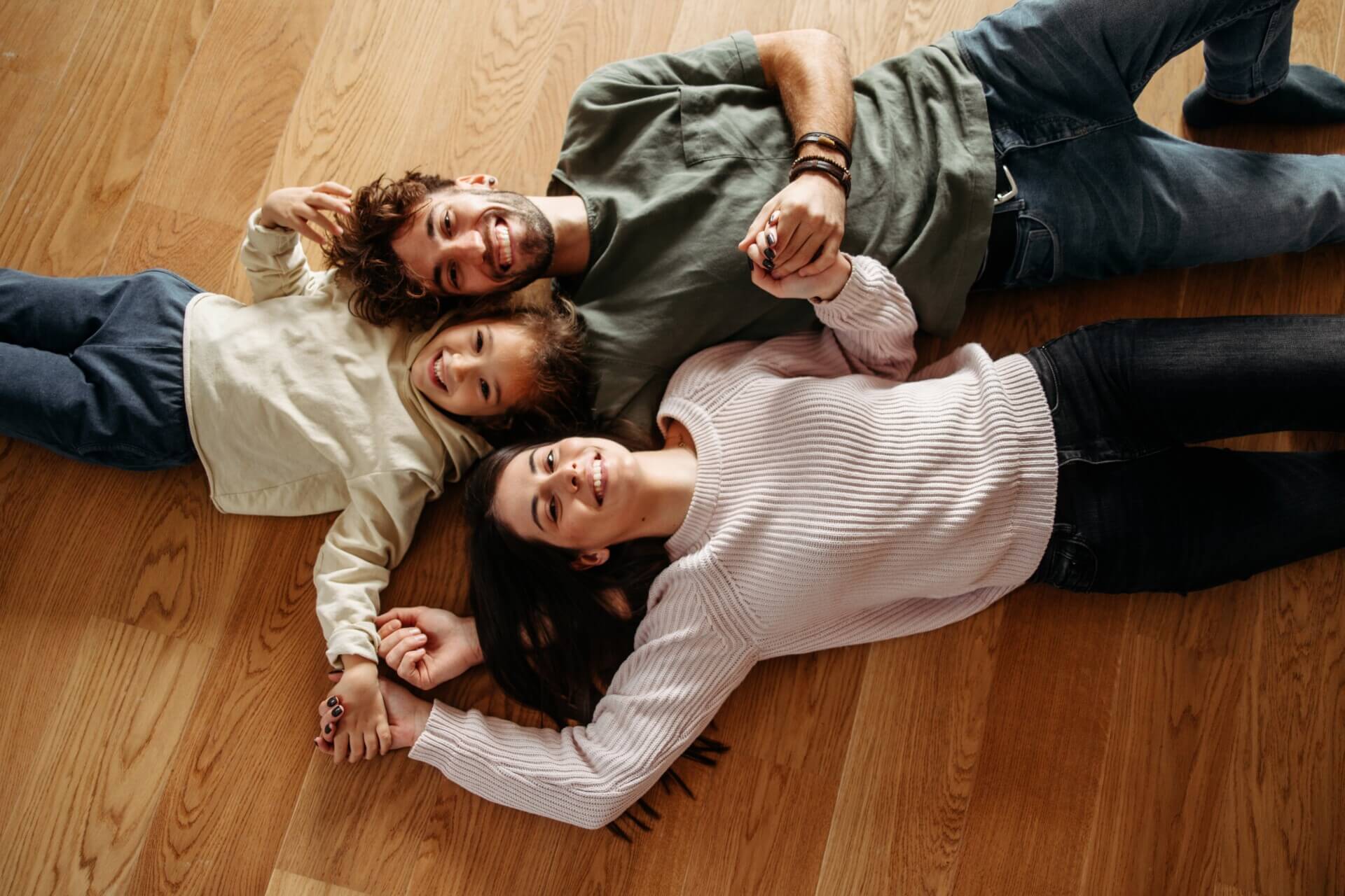 A smiling family of three lying on a hardwood floor, holding hands and looking relaxed.