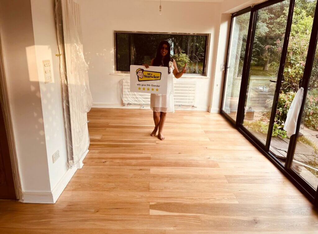 A happy customer standing barefoot on her beautifully restored wooden floor in her UK home, holding a 5-star rating card from Mr Sander®. The room is bright and inviting, with large windows allowing natural light to enhance the wood's natural grain.