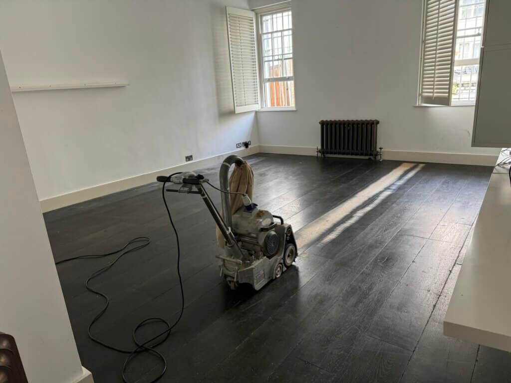 A man from Mr Sander® sanding a parquet floor in a living area in the UK using a Bona Belt sander (AMO110000.3). The machine has a drum width of 200 & 250 mm, sanding belt size 200 x 750 mm or 250 x 750 mm, motor power of 2.2 kW, and weighs 74 kg.