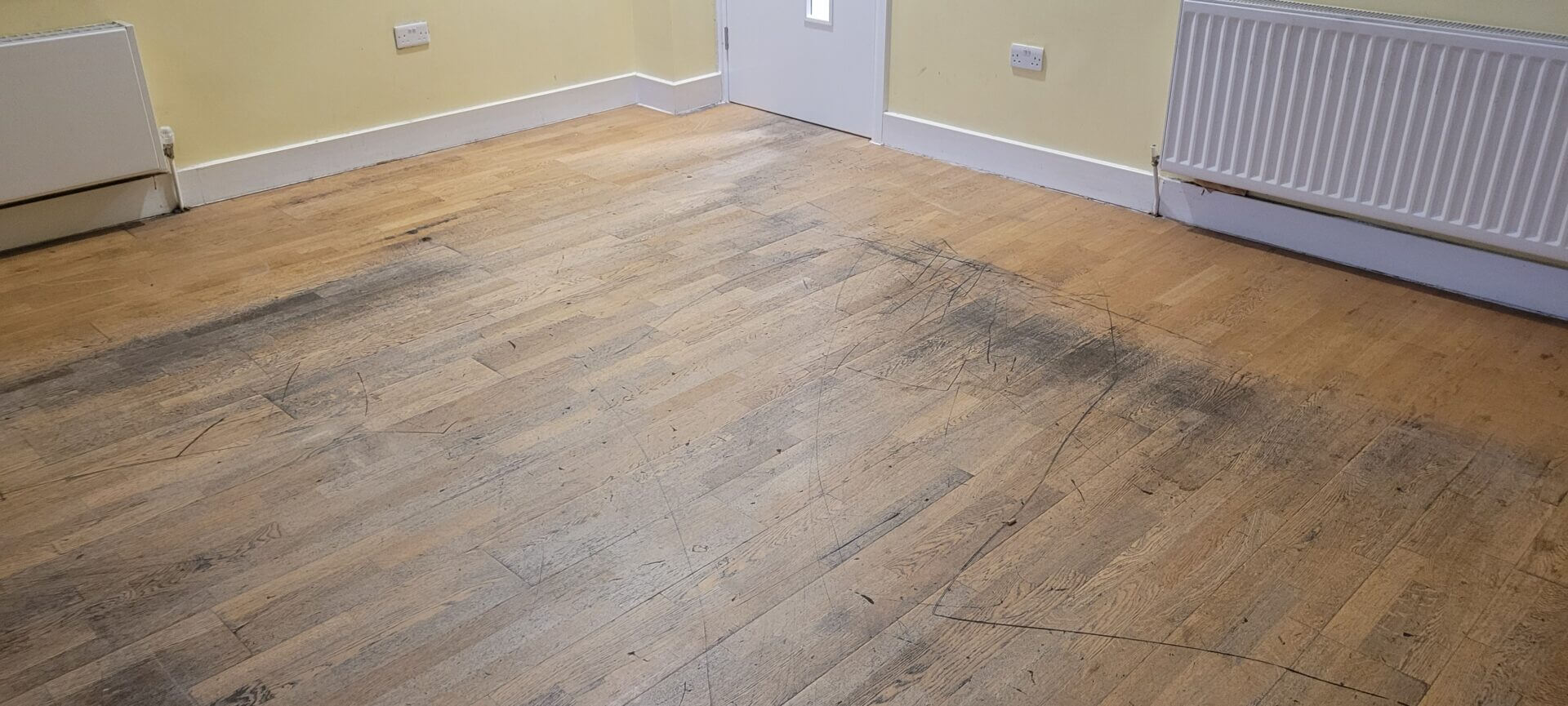 Heavily scratched wooden floor in a large room, showing scuff marks and a dull surface in need of repair.