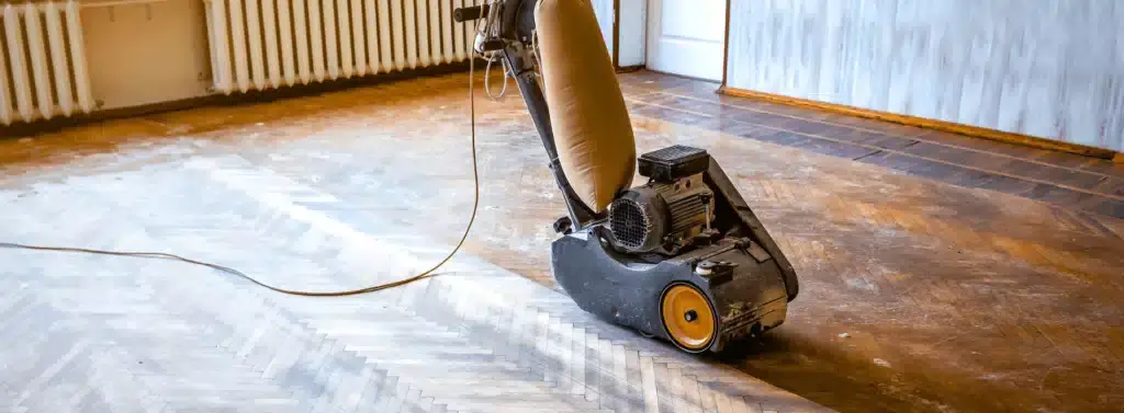 A floor sanding machine working on a herringbone parquet floor in the process of restoration. The floor displays a contrast between the sanded and unsanded sections, highlighting the transformation.