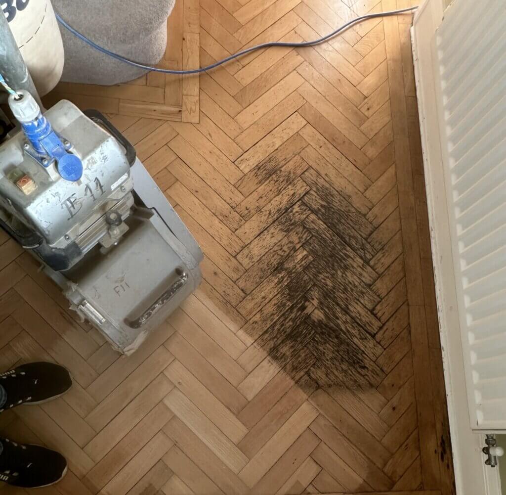 A partially sanded herringbone parquet floor with visible dark patches before restoration, demonstrating how to sand floors diagonally.