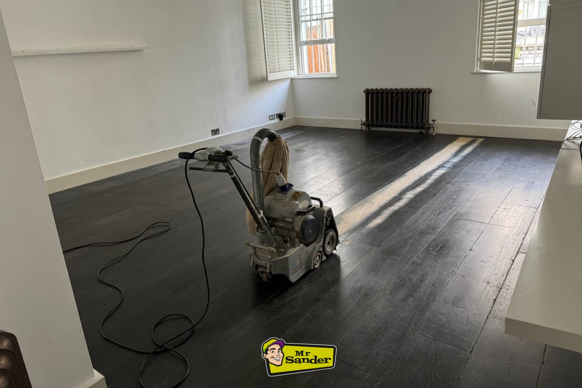 Floor sanding machine in action on dark-stained hardwood floor in Hickstead, showing floor restoration in progress with natural light streaming through windows.