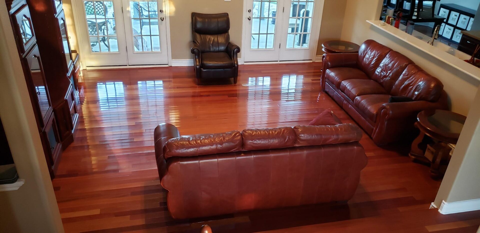 Spacious lounge with glossy, cherry‑toned Wood Stain floorboards reflecting light, furnished with leather sofas and armchair.