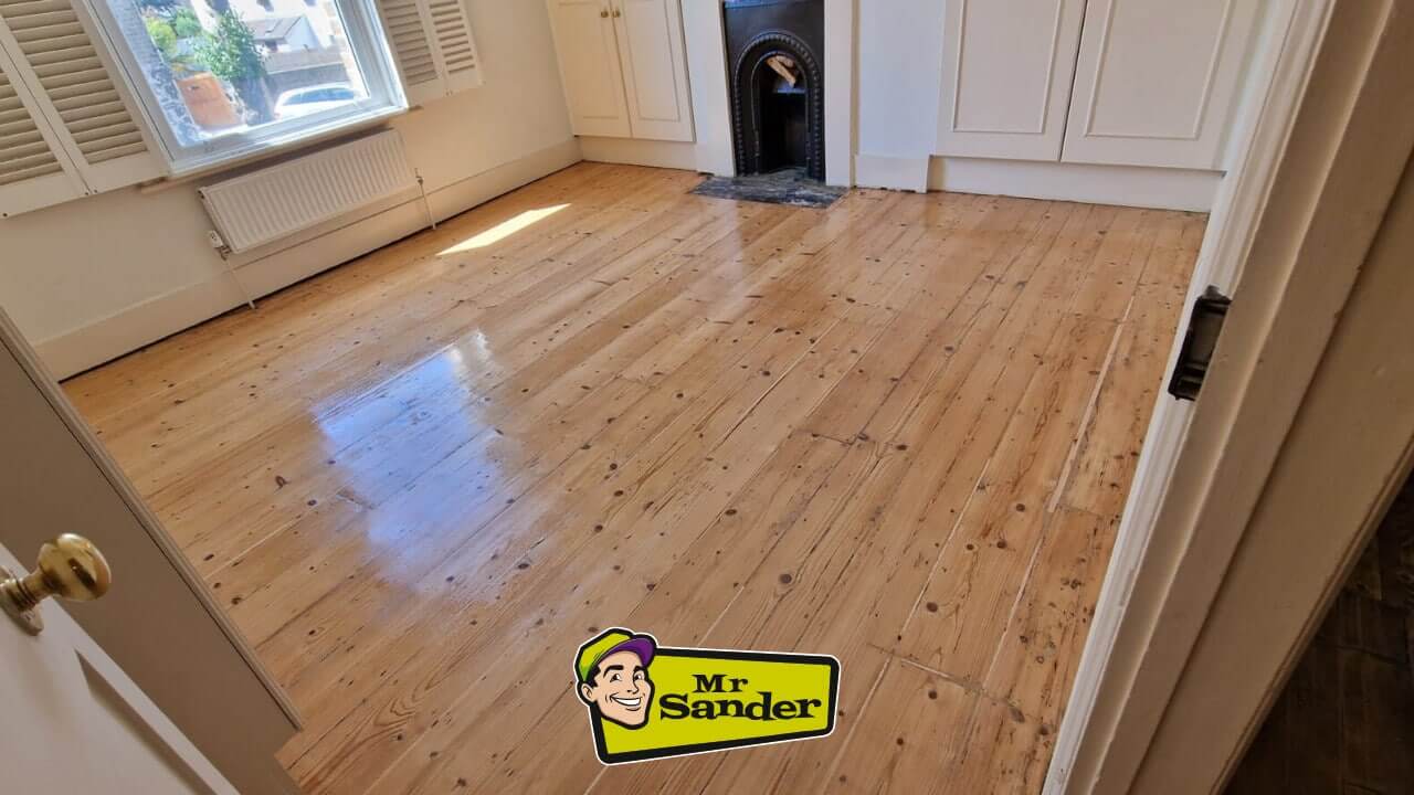 A beautifully restored wooden floor in a bright, cozy room featuring a fireplace and large window with shutters.