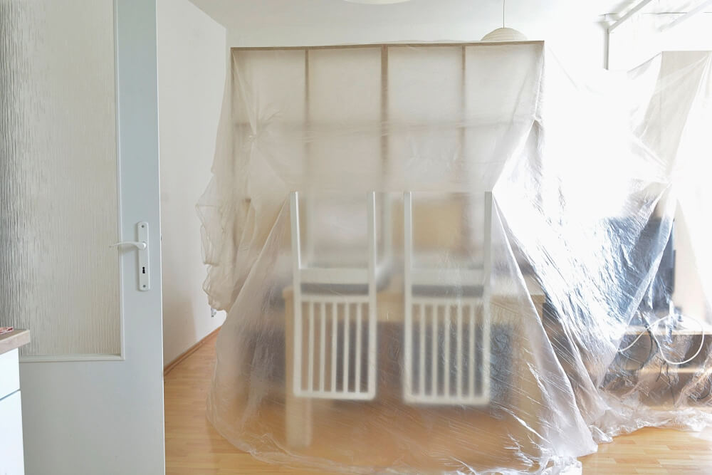 A room in preparation for floor sanding and refinishing, with furniture covered in protective plastic sheets. Two white chairs are upside down on the table, and the wooden floor is visible underneath. The room is well-lit with a large window, and cables are seen on the side. The process is being done by Mr Sander®, and the wooden floor is being protected for refinishing.