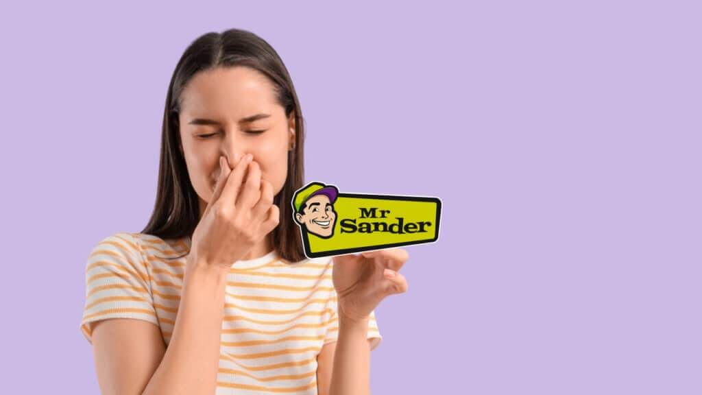 A woman pinches her nose while holding a Mr Sander® logo card against a lavender background.