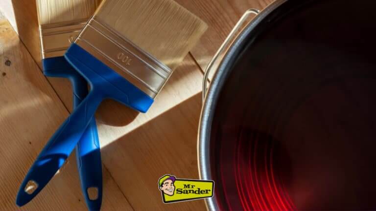 Two paintbrushes and a can of dark red paint on a wooden surface. The Mr. Sander logo is visible in the bottom right corner.