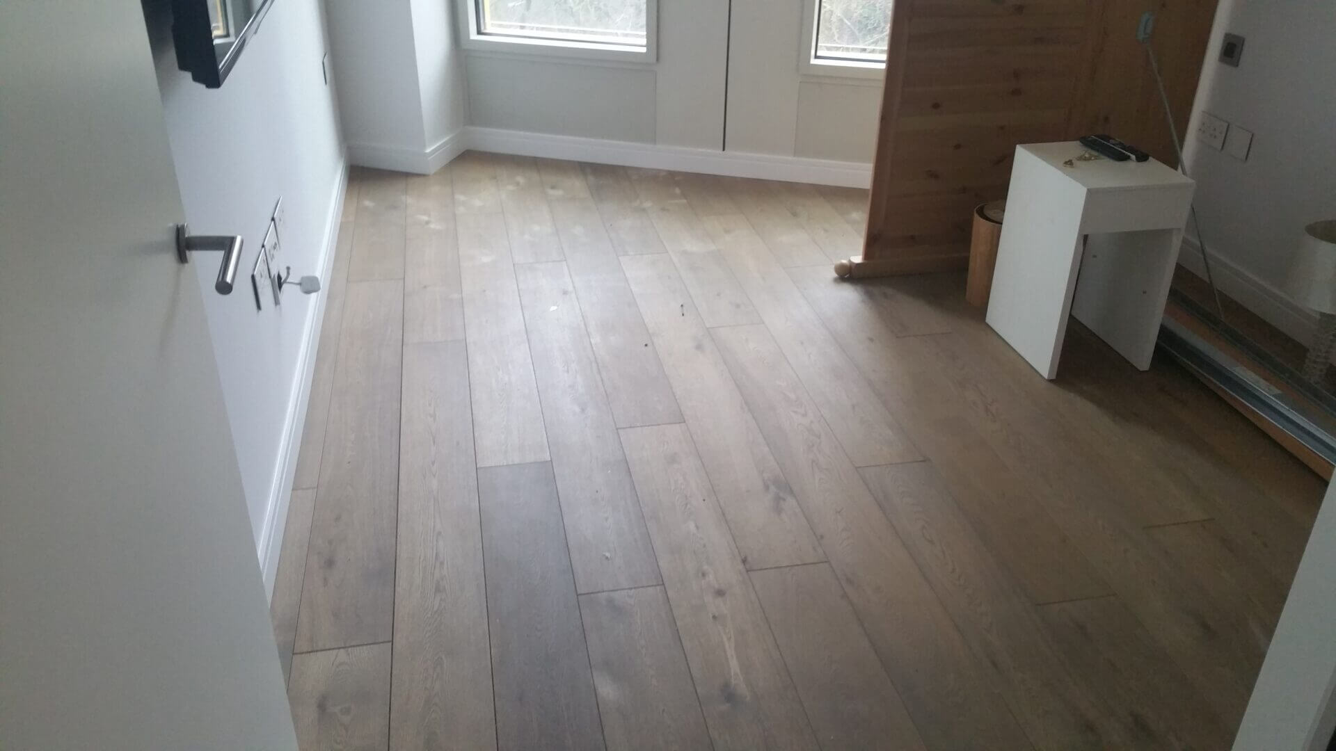 A light-toned engineered wood floor in a bright, minimal room, featuring large windows and a neutral wall palette