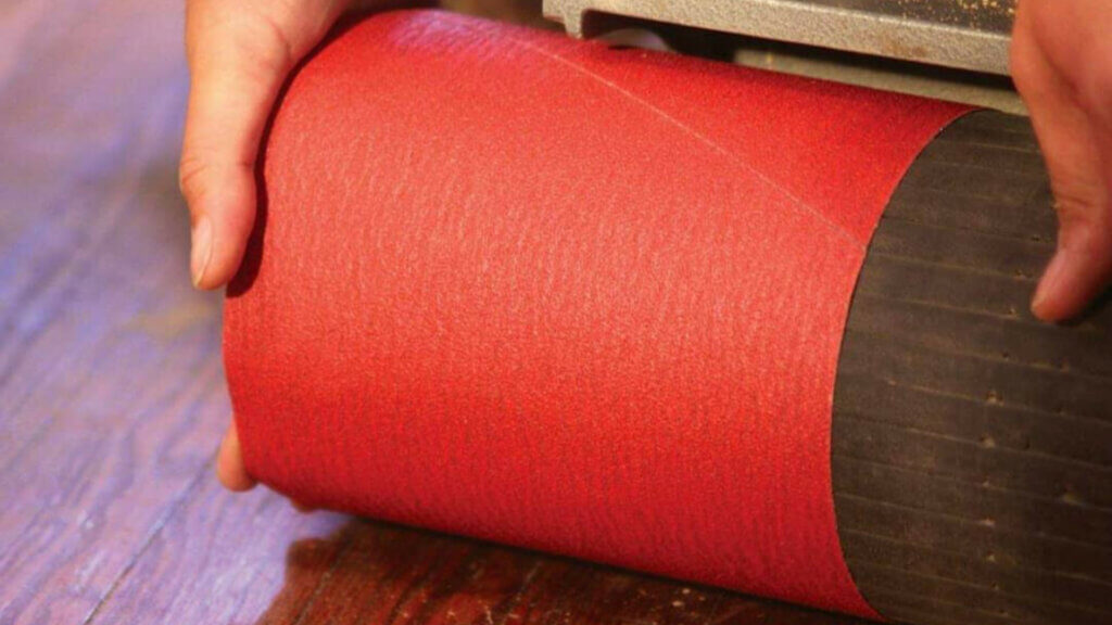 Close-up of a person's hands adjusting a roll of red sandpaper on a sanding machine. The sandpaper is being prepared for use in the floor sanding process, showing the attention to detail required in the task.