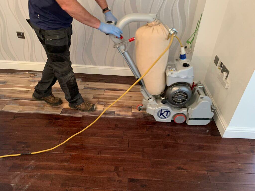 A man from Mr Sander® is sanding an oak floor in a living room in Crossness, UK, using a Bona Belt UX (AMK173100) professional belt sander. The machine specifications include a motor power of 2.2 kW, voltage of 230 V, frequency of 50 Hz, sanding belt size of 200x750 mm, drum width of 200 mm, and weight of 85 kg.
