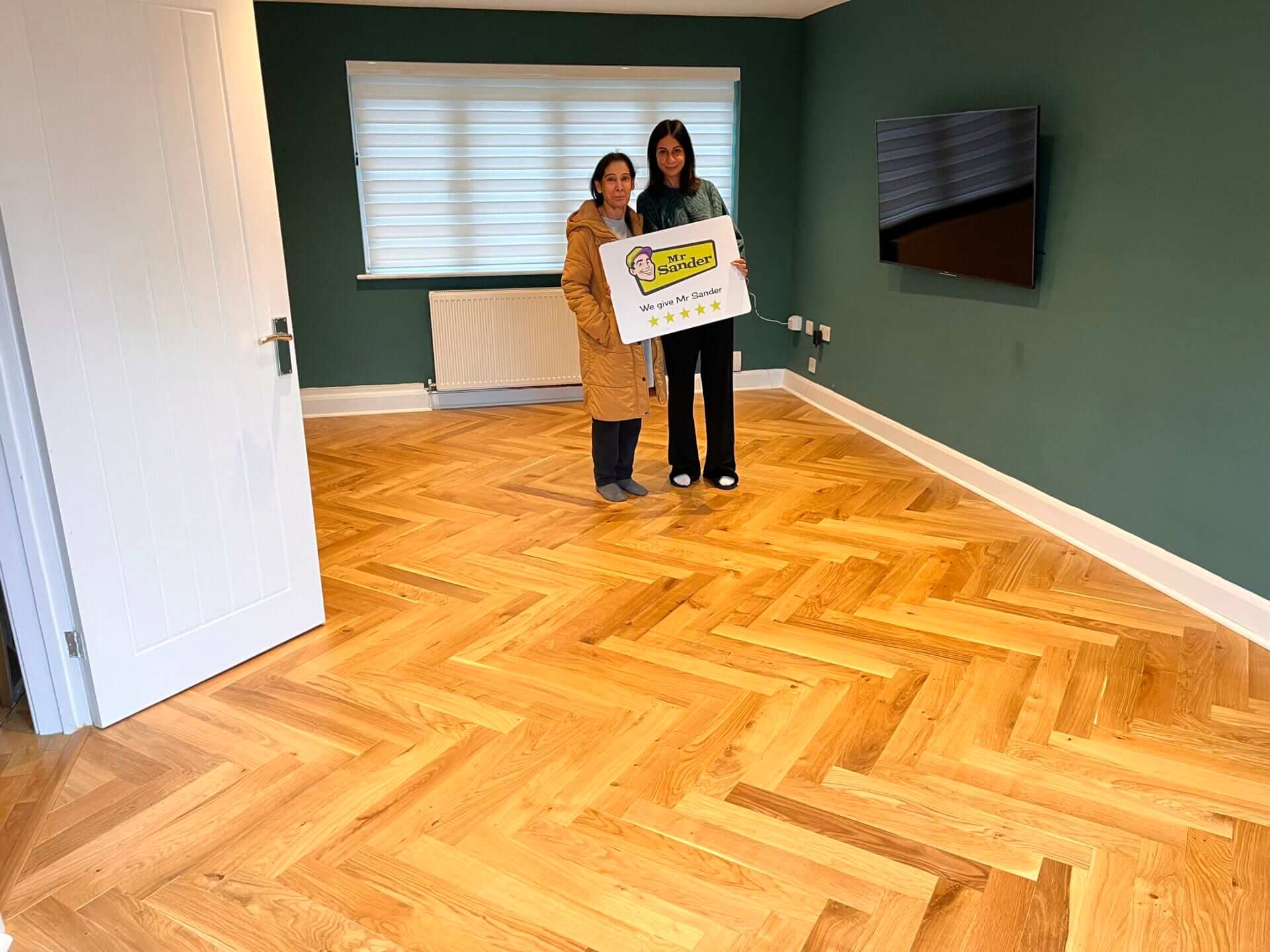 Two women standing in a modern room with freshly sanded and polished herringbone wooden flooring. They are holding a sign that reads "Mr. Sander - We give Mr. Sander 5 stars."