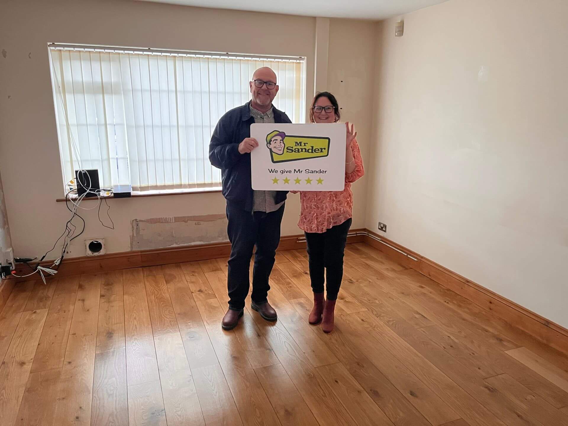 A couple standing in a room with newly sanded and refinished wooden flooring, holding a sign that reads "Mr Sander® - We give Mr Sander 5 stars." The room has light-colored walls, a large window with vertical blinds, and visible wiring on the wall.