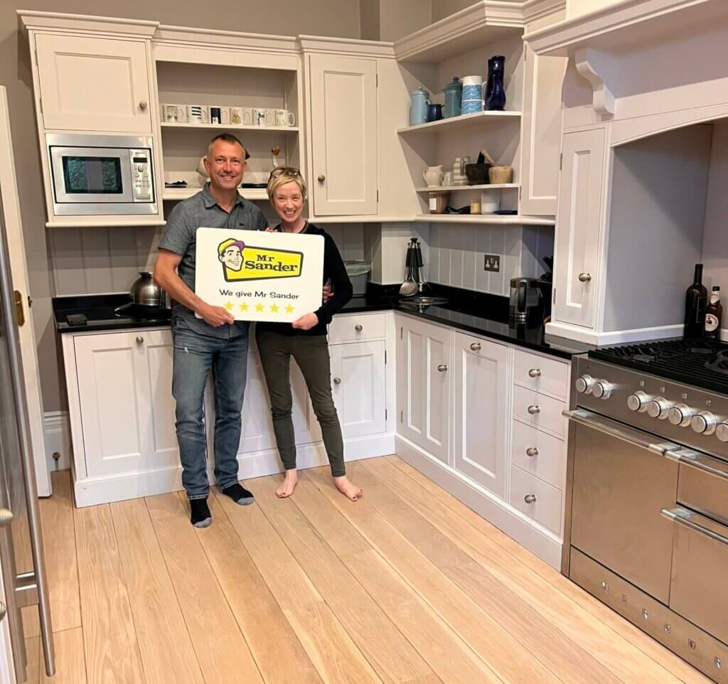 A smiling couple standing in a newly renovated kitchen with beautifully sanded and polished wooden floors. They are holding a sign that reads "Mr. Sander - We give Mr. Sander 5 stars."