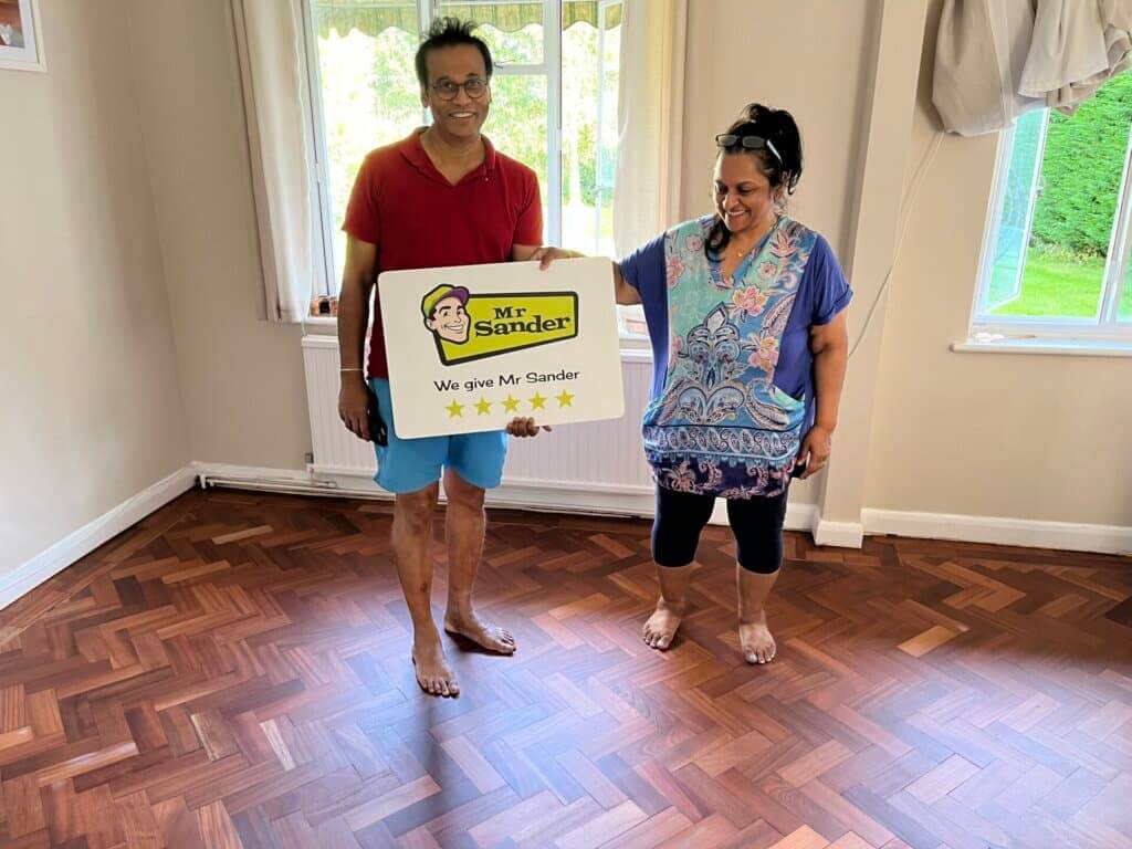 A couple happily stands on their newly refinished herringbone parquet wooden floor, holding a sign with the Mr Sander® logo that reads, "We give Mr Sander® 5 stars." The room is bright, with beige walls, a white radiator, and large windows showing a view of the garden outside. The floor's rich wood tones and intricate herringbone pattern are prominently displayed.