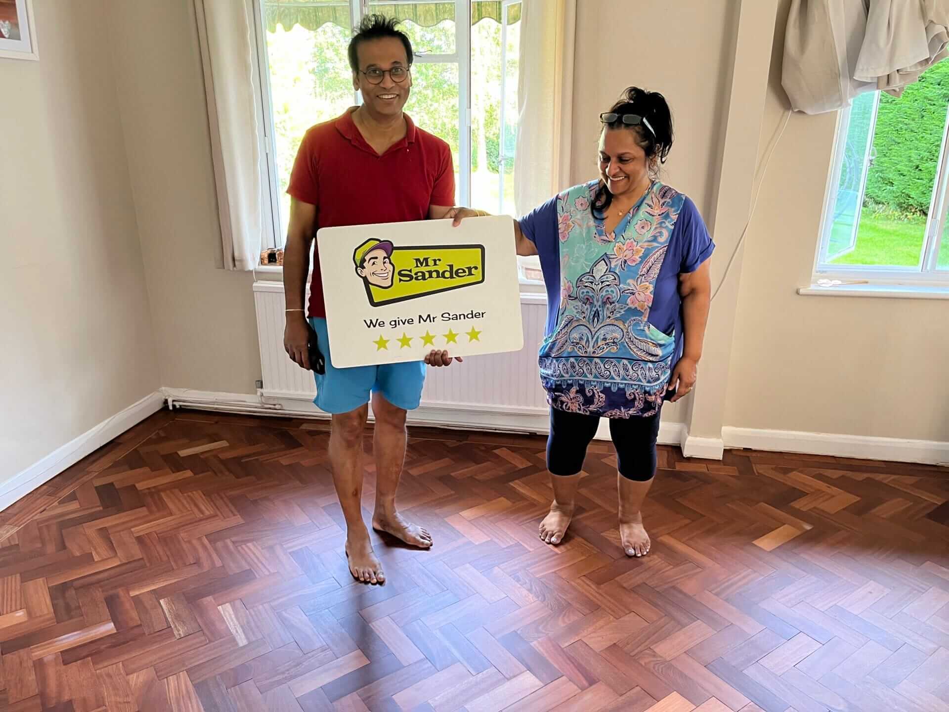 A couple happily stands on their newly refinished herringbone parquet wooden floor, holding a sign with the Mr Sander® logo that reads, "We give Mr Sander® 5 stars." The room is bright, with beige walls, a white radiator, and large windows showing a view of the garden outside. The floor's rich wood tones and intricate herringbone pattern are prominently displayed.