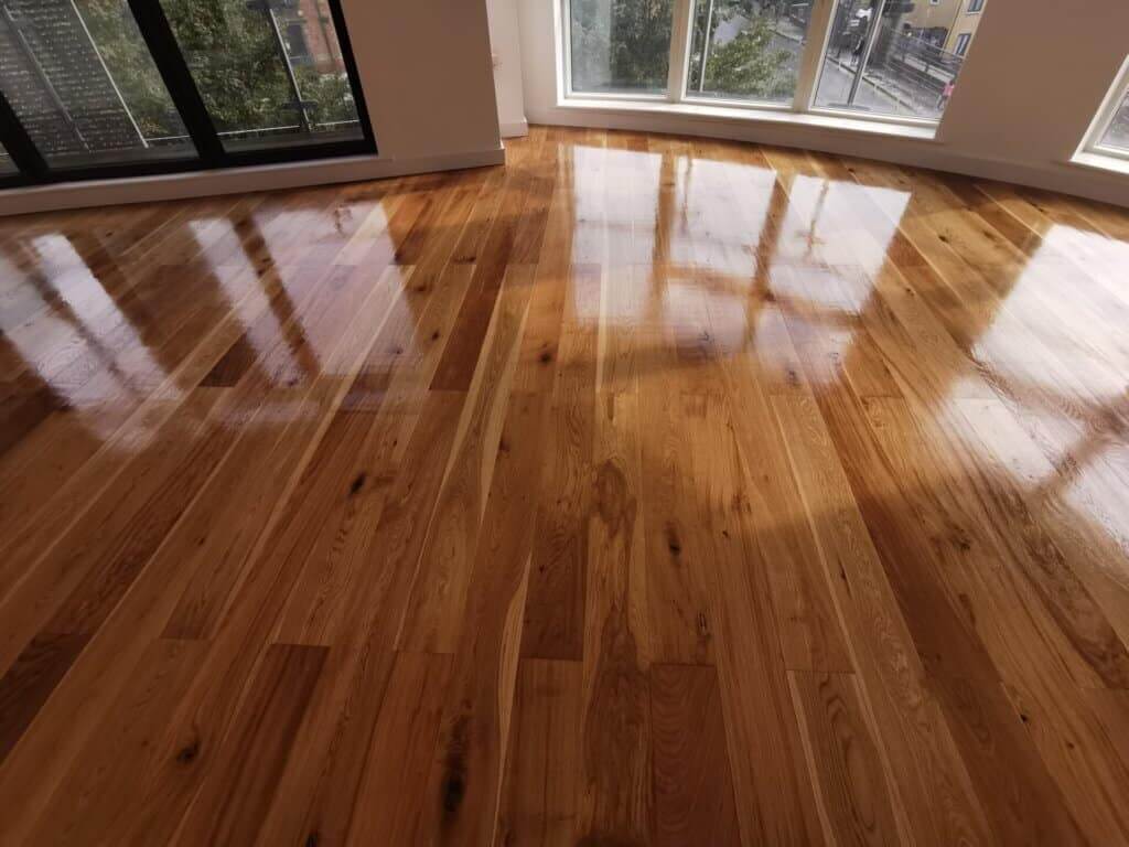 A beautifully refinished oak hardwood floor showcases a glossy finish, reflecting natural light from large windows. The floor exhibits a rich grain pattern and a polished surface, emphasizing the quality of craftsmanship achieved through the sanding and refinishing process by Mr. Sander®. The bright and airy space highlights the elegance of the newly restored flooring.