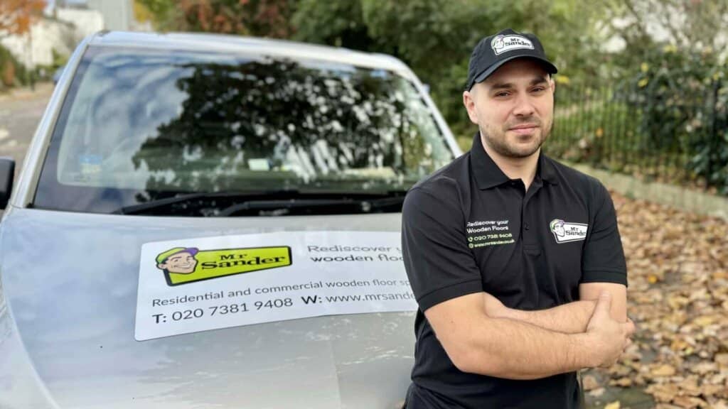 A man from Mr Sander® stands with his arms crossed in front of a company vehicle. He is wearing a black polo shirt and cap with the Mr Sander® logo. The vehicle features a sign with the company's logo and contact information, including the phone number 020 7381 9408 and the website www.mrsander.com. The background shows a leafy residential street.