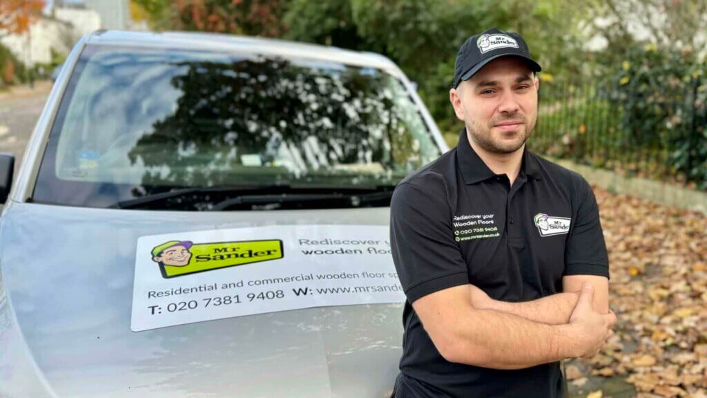 A professional from Mr Sander® stands confidently with arms crossed in front of a company vehicle branded with the Mr Sander® logo and contact information. The image underscores the expertise and reliability of Mr Sander® in parquet flooring restoration.