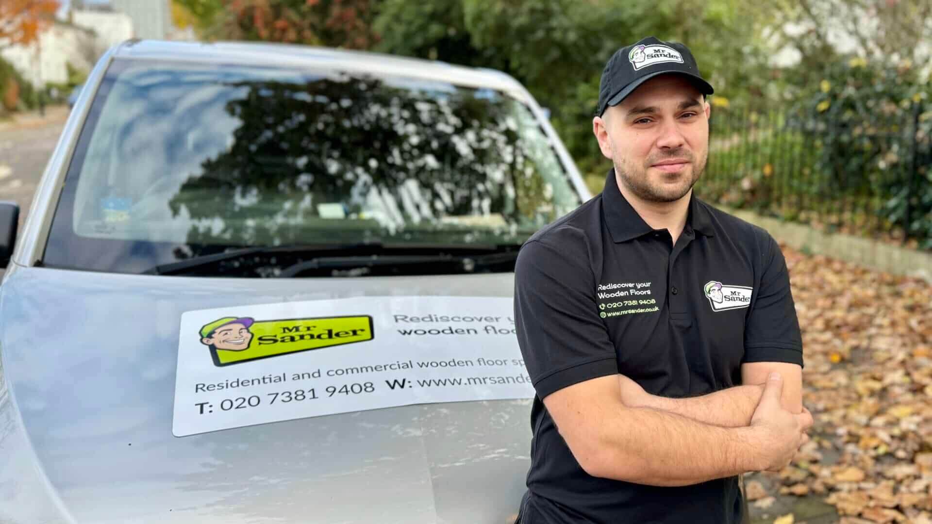 A Mr Sander® professional standing confidently in front of a company van with the Mr Sander® logo and contact information displayed. The professional is wearing a black uniform and cap with the Mr Sander® branding.