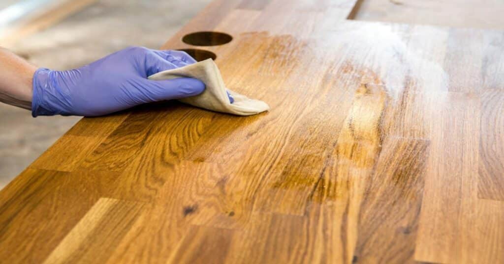 A hand in a blue glove applying oil finish to a wooden table with a cloth.