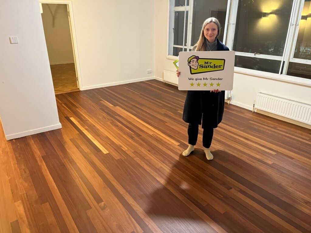A satisfied customer in New Addington, UK, standing in a room with beautifully refinished teak hardwood flooring, holding a sign that reads, "We give Mr Sander 5 stars." The image showcases the rich tones and smooth finish of the teak wood, highlighting the exceptional floor sanding and refinishing services provided by Mr Sander®. The room's large windows and minimalist decor complement the elegant flooring.
