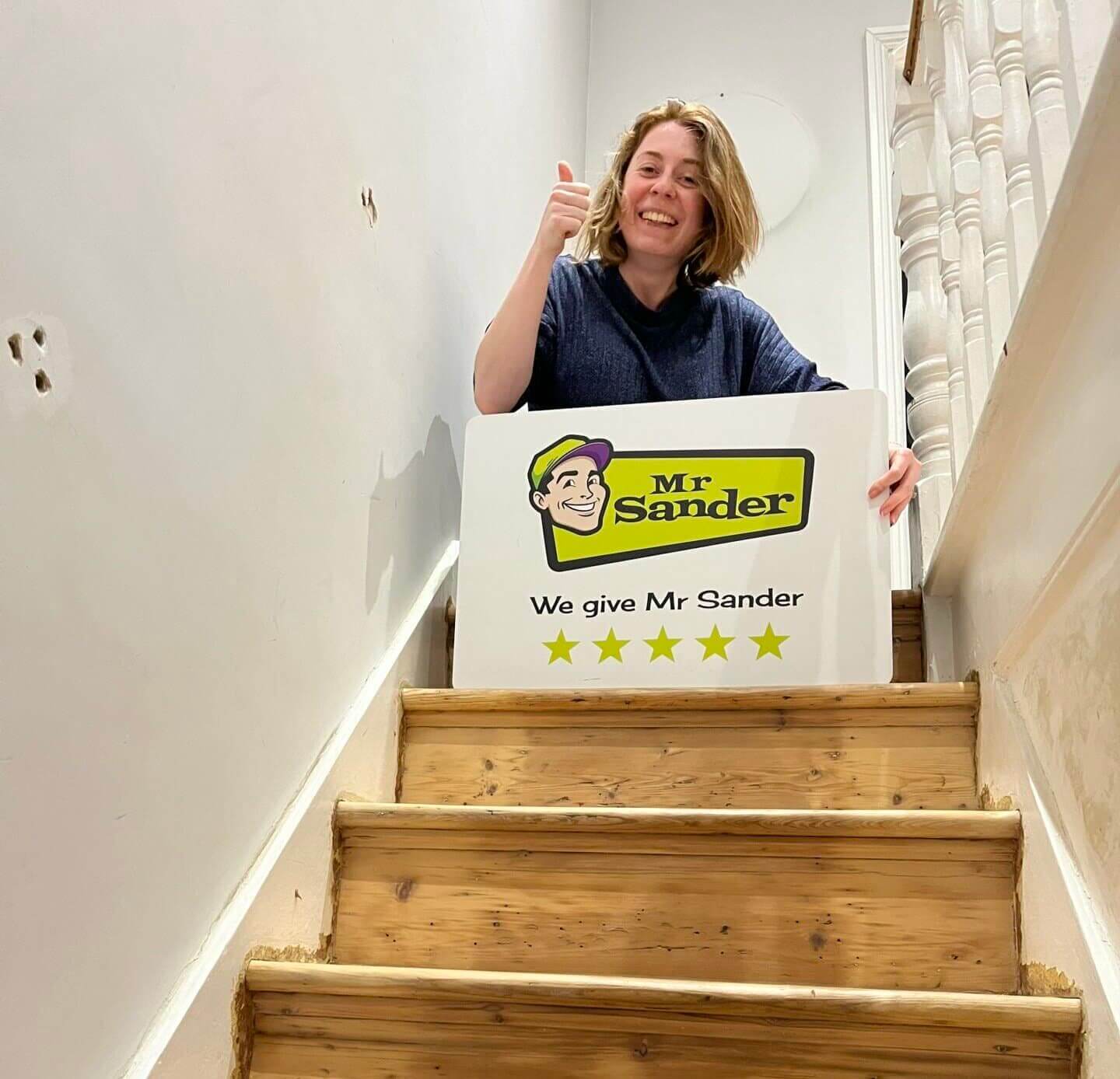 A woman giving a thumbs-up and holding a sign that reads "Mr. Sander - We give Mr. Sander 5 stars," standing on a staircase with newly sanded wooden steps.
