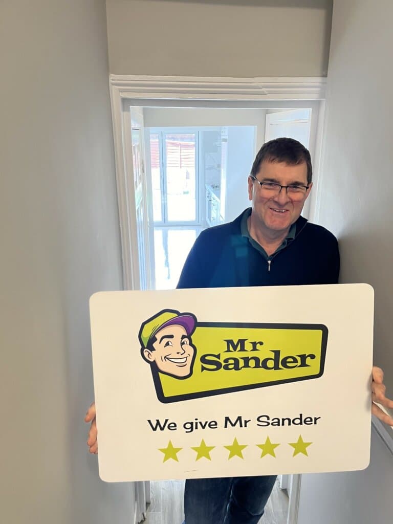 A satisfied customer holding a sign that reads "Mr Sander® - We give Mr Sander 5 stars" in a freshly refinished room with sanded and polished wooden floors.