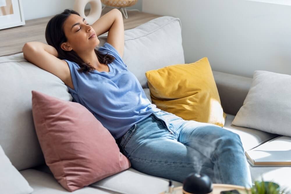 A woman relaxing on a beige sofa with colorful cushions, including pink and yellow. She appears peaceful, leaning back with her arms resting behind her head, enjoying the comfort of a cozy living room with natural light. The flooring beneath is a beautifully refinished oak hardwood floor.