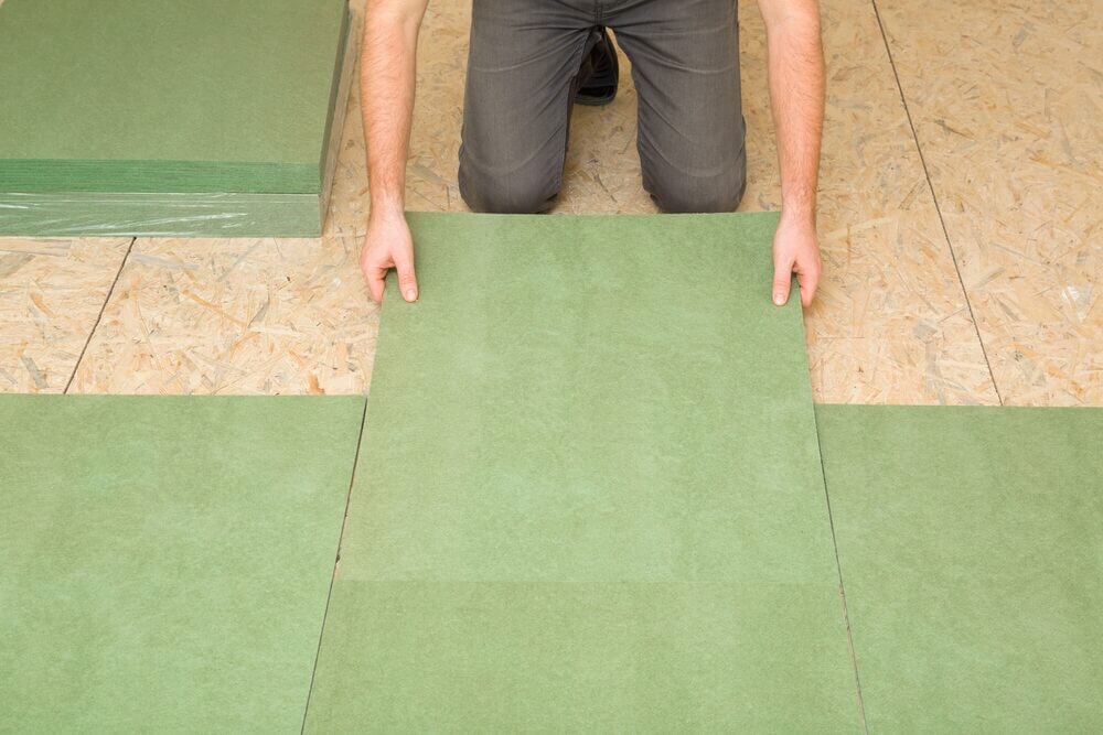 A floor installer kneels on an OSB subfloor, carefully positioning green underlay panels to prepare for new flooring.