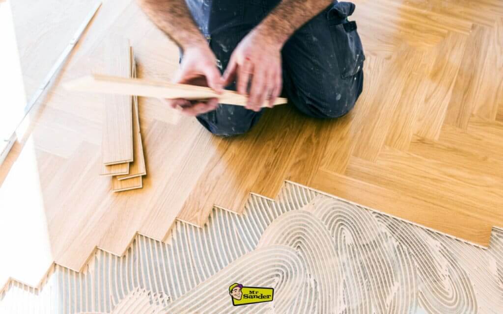 A person kneeling on a wood subfloor, carefully fitting herringbone-style parquet boards into fresh adhesive.