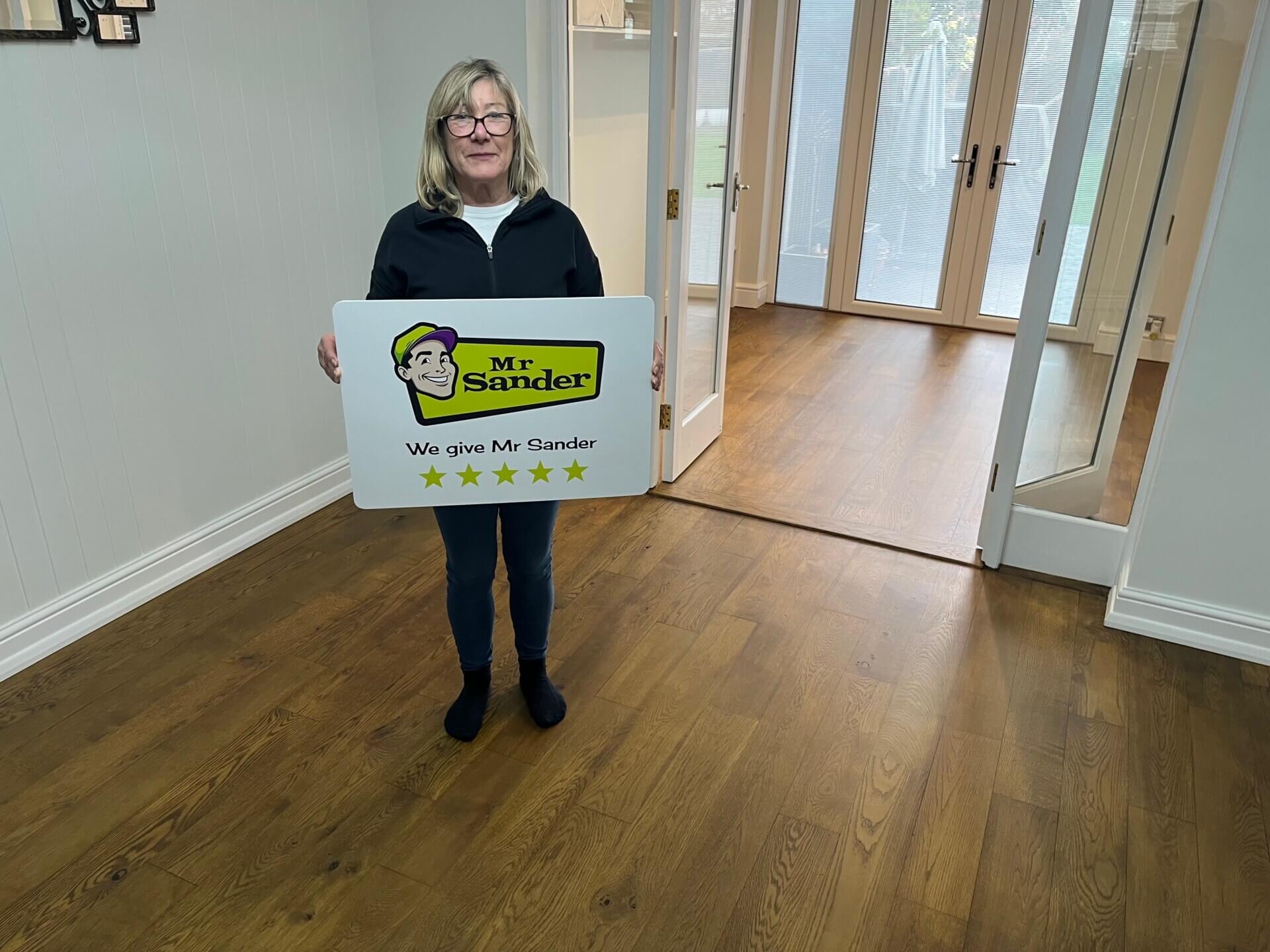 A satisfied customer standing in a room with newly refinished hardwood floors in Harpenden, UK, holding a sign that reads "Mr Sander® - We give Mr Sander 5 stars". The room features large glass doors and ample natural light.