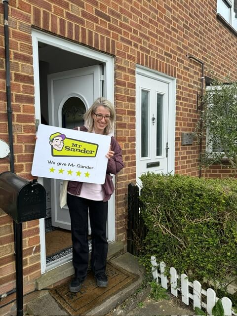 A woman stands at the entrance of a brick house, holding a sign that reads "Mr Sander - We give Mr Sander ★★★★★". The house features a white front door and a small garden with a hedge and a white picket fence.