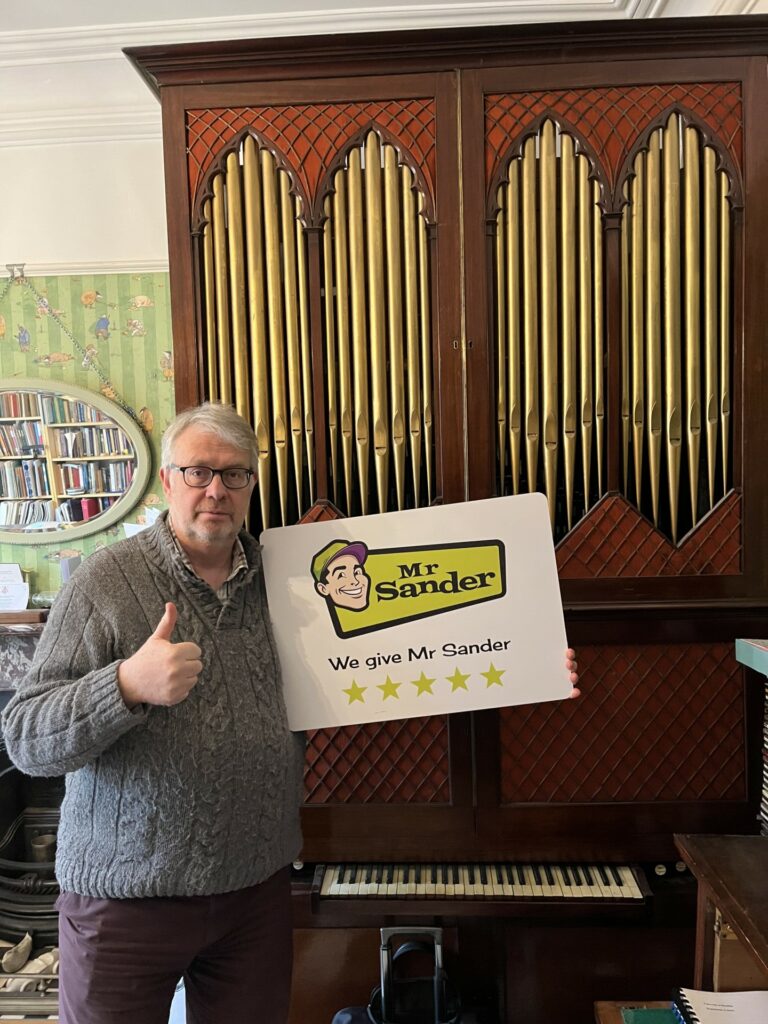 A man wearing glasses and a gray sweater stands in front of a vintage organ, holding a sign that reads "Mr. Sander - We give Mr. Sander 5 stars." The background features a bookshelf and a decorative mirror. The floor sanding and refinishing were completed by Mr. Sander®, UK, on a wooden parquet floor.