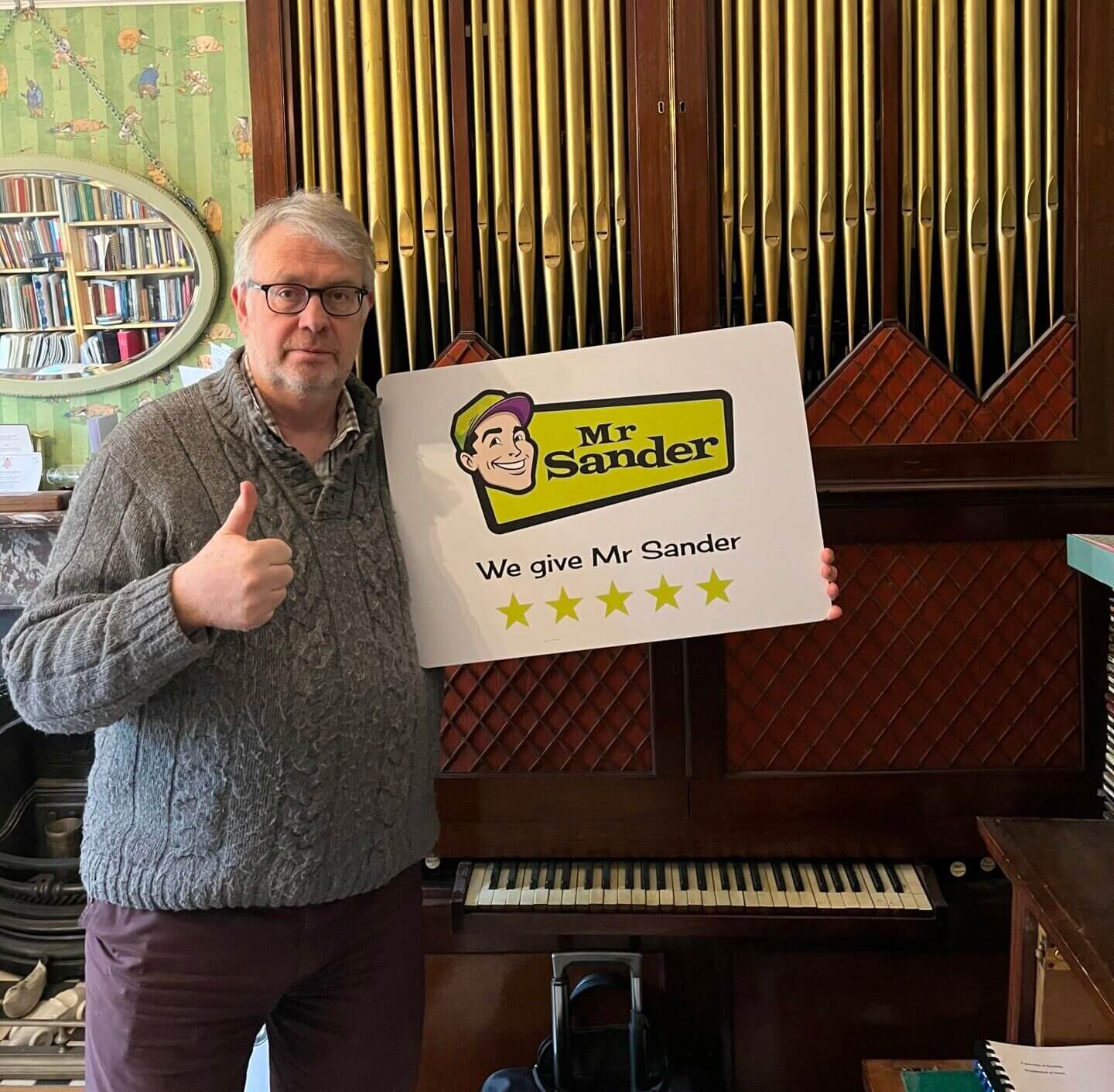A man wearing glasses and a gray sweater stands in front of a vintage organ, holding a sign that reads "Mr. Sander - We give Mr. Sander 5 stars." The background features a bookshelf and a decorative mirror. The floor sanding and refinishing were completed by Mr. Sander®, UK, on a wooden parquet floor.