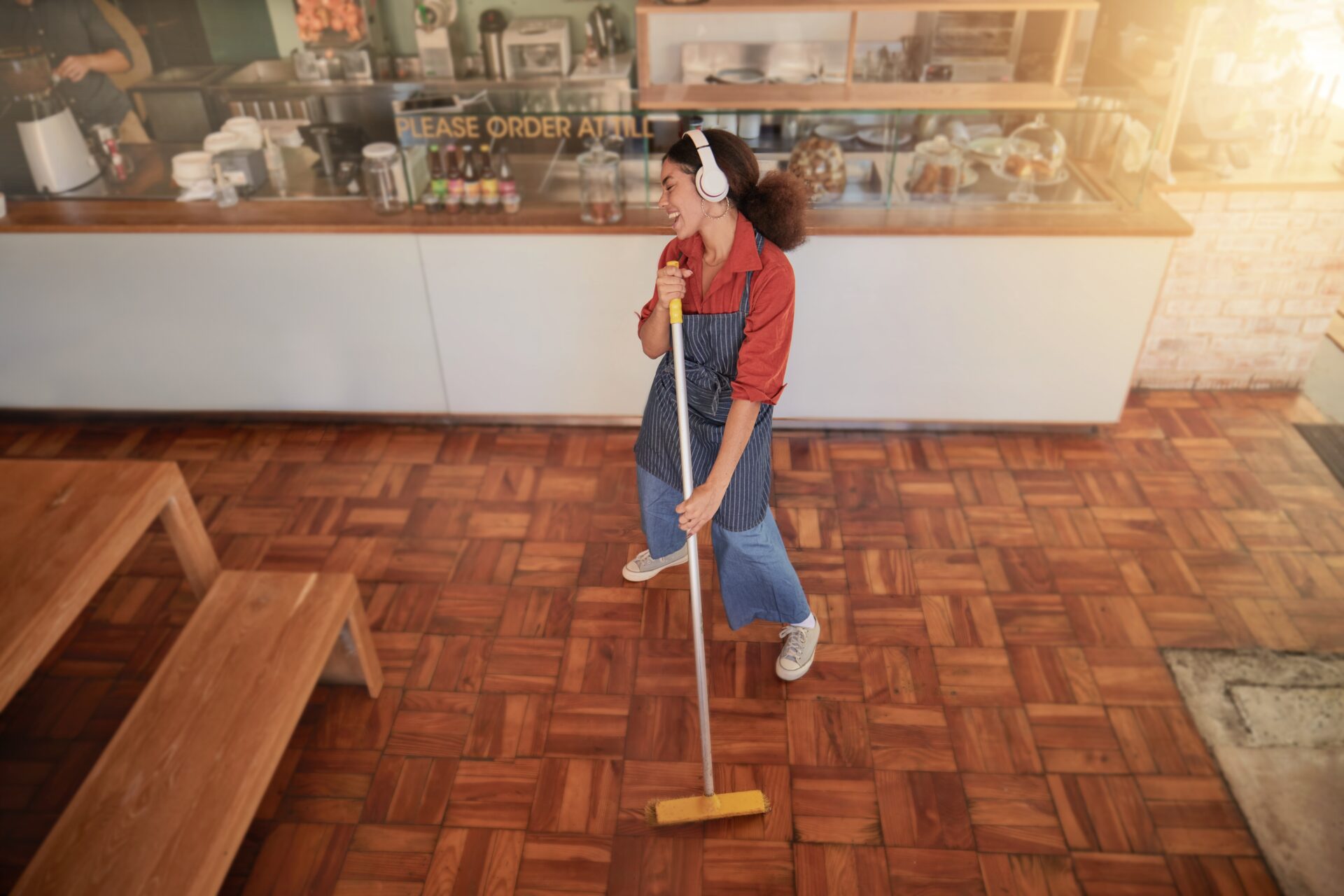 Young bakery worker wearing headphones sweeps a parquet floor installed for bakeries, showing easy-clean wood blocks after service.