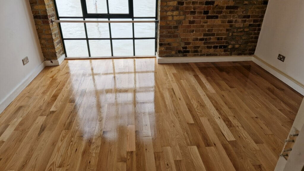 A newly lacquered wooden floor in a room with a rustic brick wall and a large window overlooking a body of water. The floor has a shiny finish, reflecting the light and the outdoor scenery. The room has white walls and minimalist decor, showcasing the natural beauty of the polished wood.