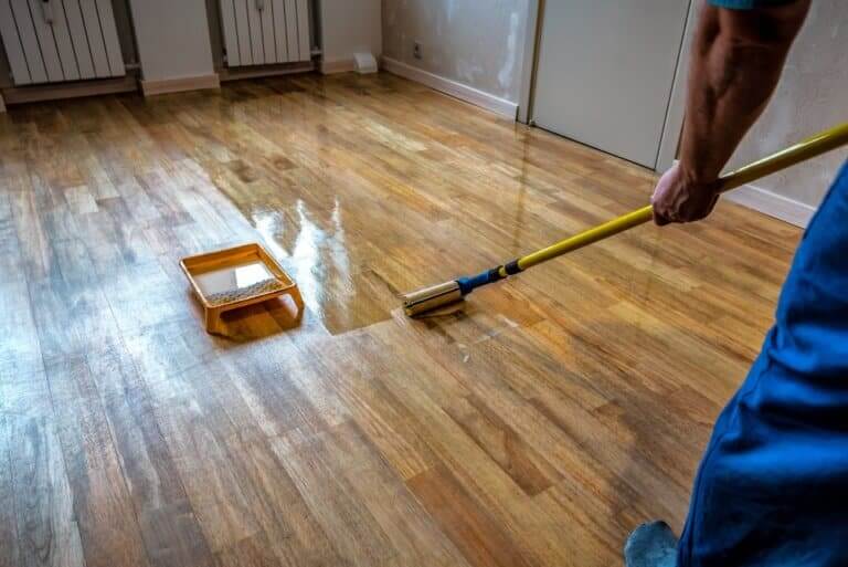 A person is applying a lacquer finish to a wooden floor using a wide applicator brush attached to a long handle. The floor has a glossy appearance where the finish has been applied, contrasting with the unfinished section. A tray filled with lacquer is placed on the floor, and the person is wearing blue work pants.