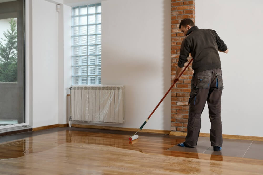A worker is applying a glossy finish to a wooden floor using a roller with a long handle. The room features a brick pillar, large windows, and a radiator covered with protective plastic. The unfinished floor contrasts with the newly coated section, which reflects light and has a smooth, polished appearance.