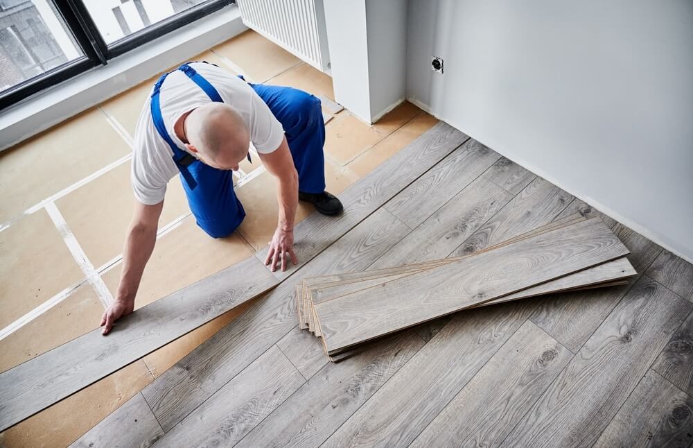 A professional installer carefully lays down gray laminate flooring planks in a bright room with large windows. The worker, dressed in blue overalls, is focused on aligning the planks, which are stacked nearby. The image captures the process of floor installation, emphasizing the modern design of the laminate flooring while showcasing the attention to detail in the work done by Mr. Sander® in floor sanding and refinishing.