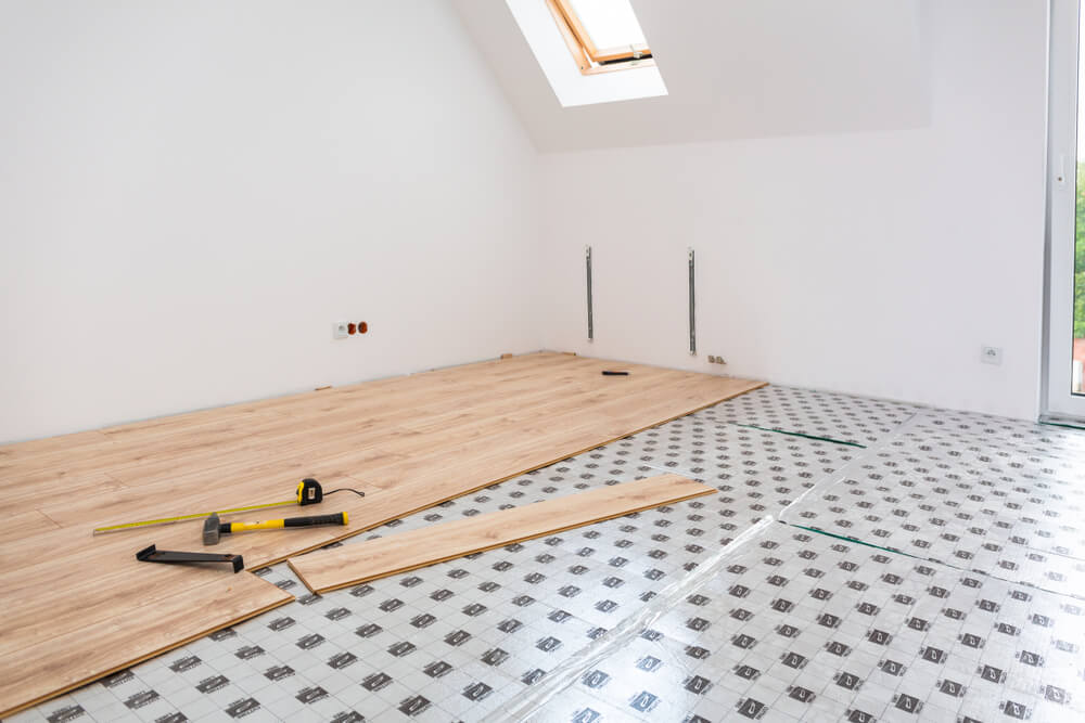 Light Oak laminate flooring boards being laid over underlayment in a bright attic, showcasing tools during a wood floor installation.