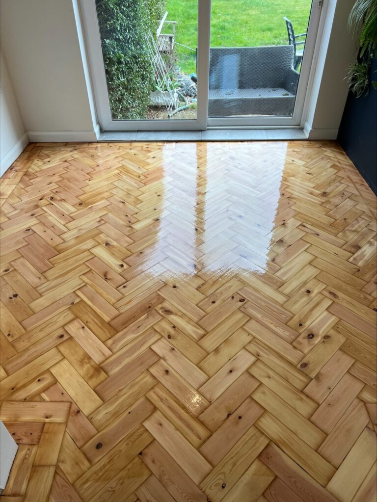 Bright, newly refinished reclaimed parquet floor in a herringbone pattern, positioned near glass doors opening onto a garden.