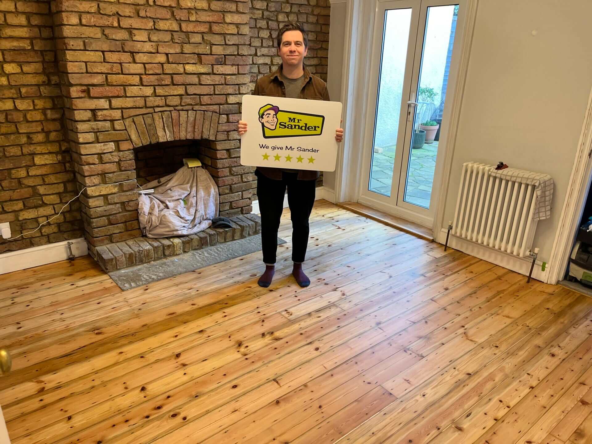 A person standing in a room with newly sanded and refinished wooden floors, holding a sign with the logo "Mr Sander®" and the text "We give Mr Sander 5 stars." A brick fireplace and large windows are in the background.