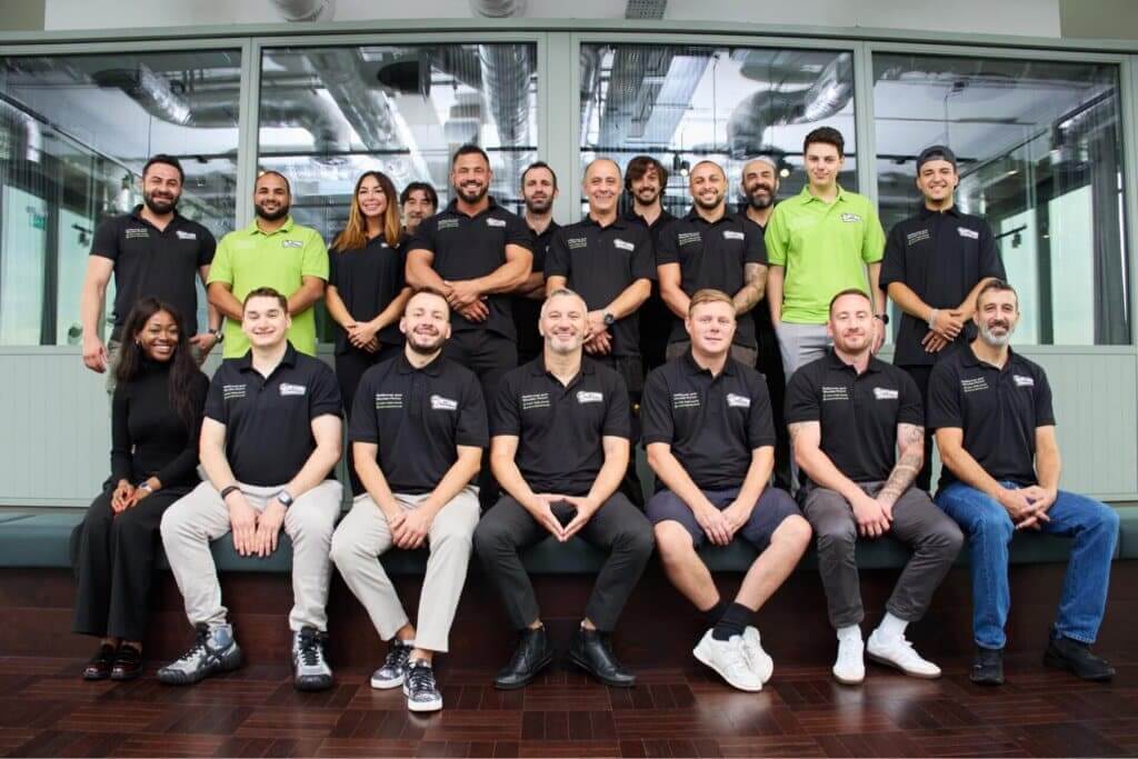 A group photo of the Mr Sander® team, specializing in floor sanding and refinishing. The diverse team is dressed in black polo shirts with the company logo, while one member stands out in a bright green shirt. They are posed together in a modern office setting, showcasing their camaraderie and professionalism in providing flooring services.