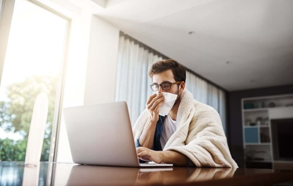 A young man wrapped in a blanket working on his laptop, pausing to blow his nose, suggesting possible respiratory or allergy-related discomfort.