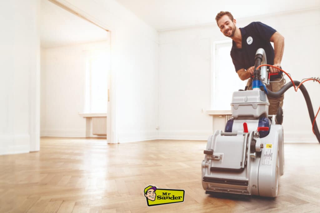 Professional floor sanding in action, featuring a smiling Mr Sander technician using a sanding machine on a light oak herringbone parquet floor.