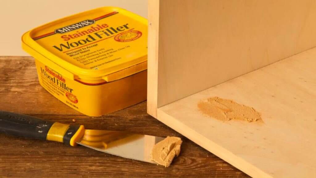 A container of Minwax Stainable Wood Filler placed on a wooden surface alongside a putty knife. A light wooden floor panel is visible with wood filler applied to a section, showcasing floor sanding and refinishing work.