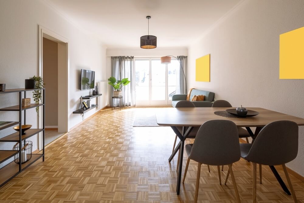 A bright and airy living and dining space with a beautifully refinished basketweave-pattern parquet wood floor. The room is furnished with a modern dining table and chairs, a cozy seating area with a sofa and coffee table, and indoor plants. Large windows allow natural light to fill the space, and minimalist decor completes the look.