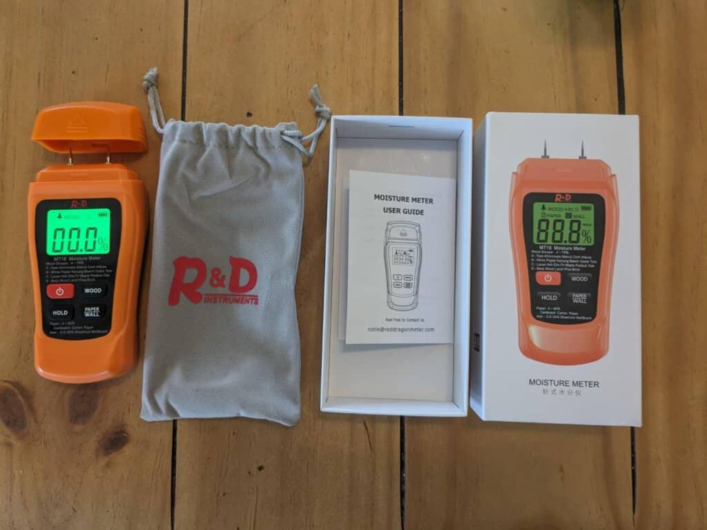 An orange moisture meter by R&D Instruments displayed with its packaging, user guide, and protective pouch on a wooden surface. The device is designed for measuring moisture levels in wood and other materials.