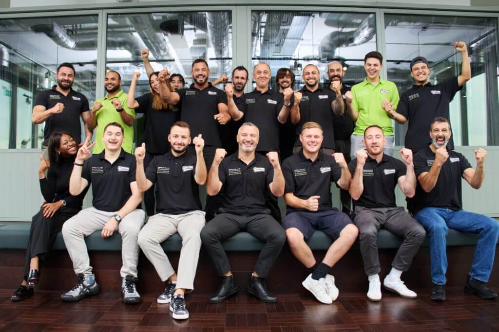 The enthusiastic Mr. Sander® team poses together, wearing black and green branded shirts, showing their excitement and professionalism. The team is gathered in front of large windows, sitting on a bench, and cheering. The floor beneath them is a dark, polished hardwood, reflecting their expertise in floor refinishing.